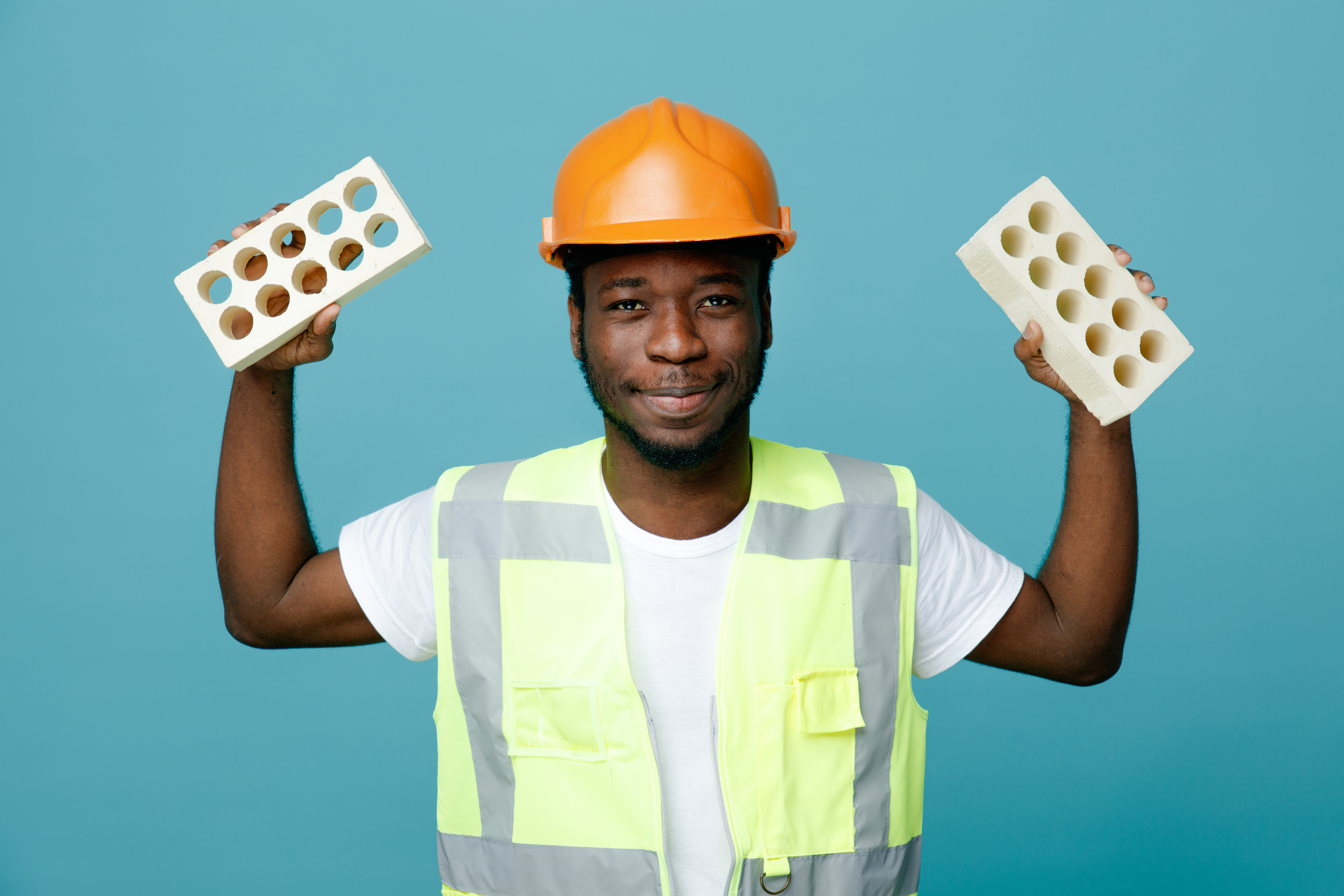 pleased young african american builder in uniform holding bricks isolated on blue background
